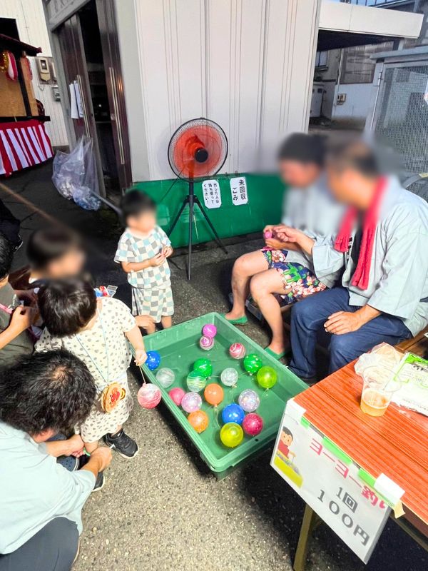 水に浮かんだヨーヨーを釣り上げようとする子どもたち。ヨーヨー釣り1回100円の看板
